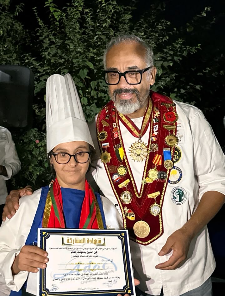 A trainee chef receiving her participation certificate alongside a decorated master chef